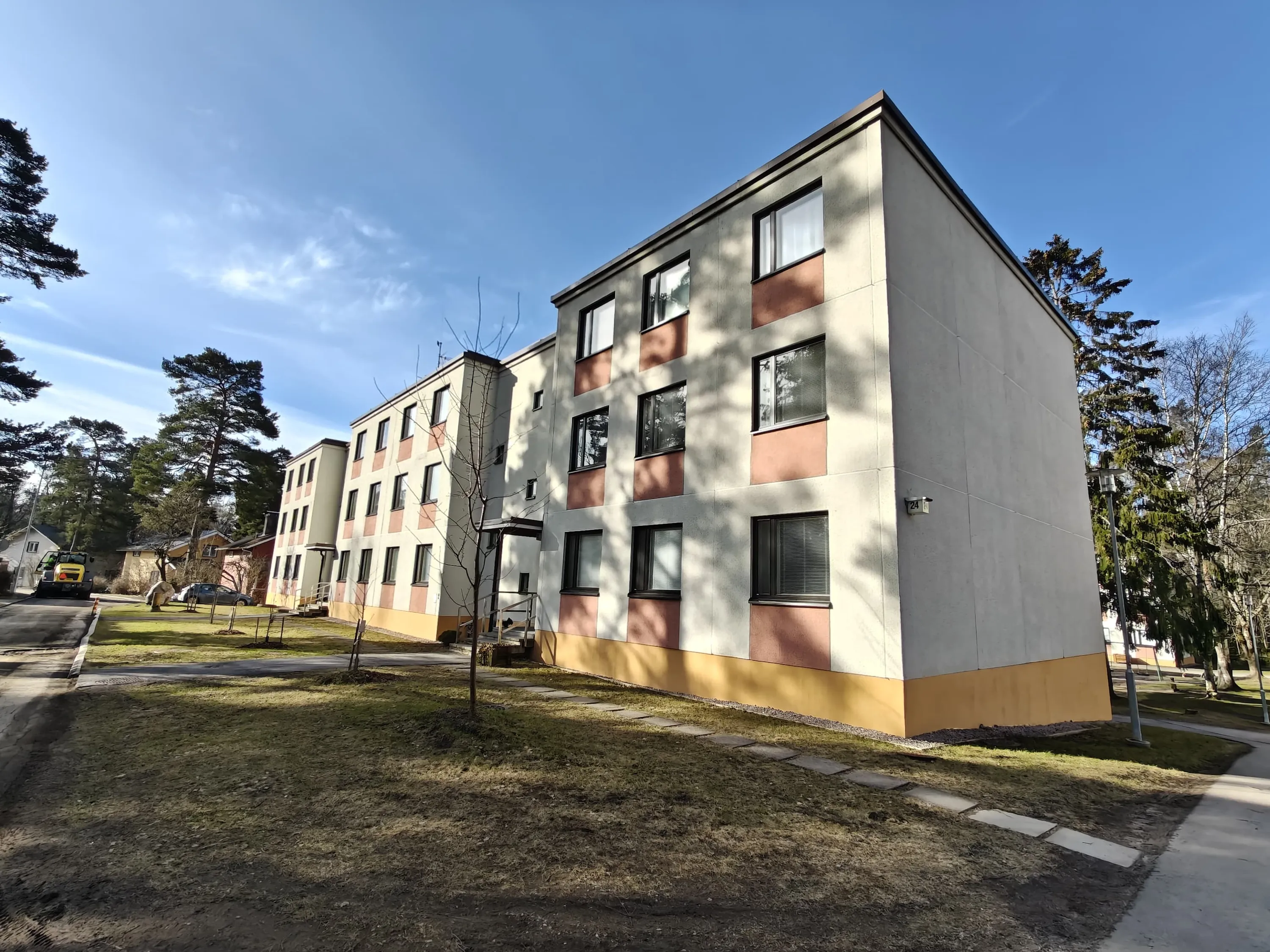Kolmikerroksinen asuintalo, jossa on harmaa julkisivu, ruskeat ikkunoiden välit ja keltainen pohjaosa, aurinkoisessa säässä.