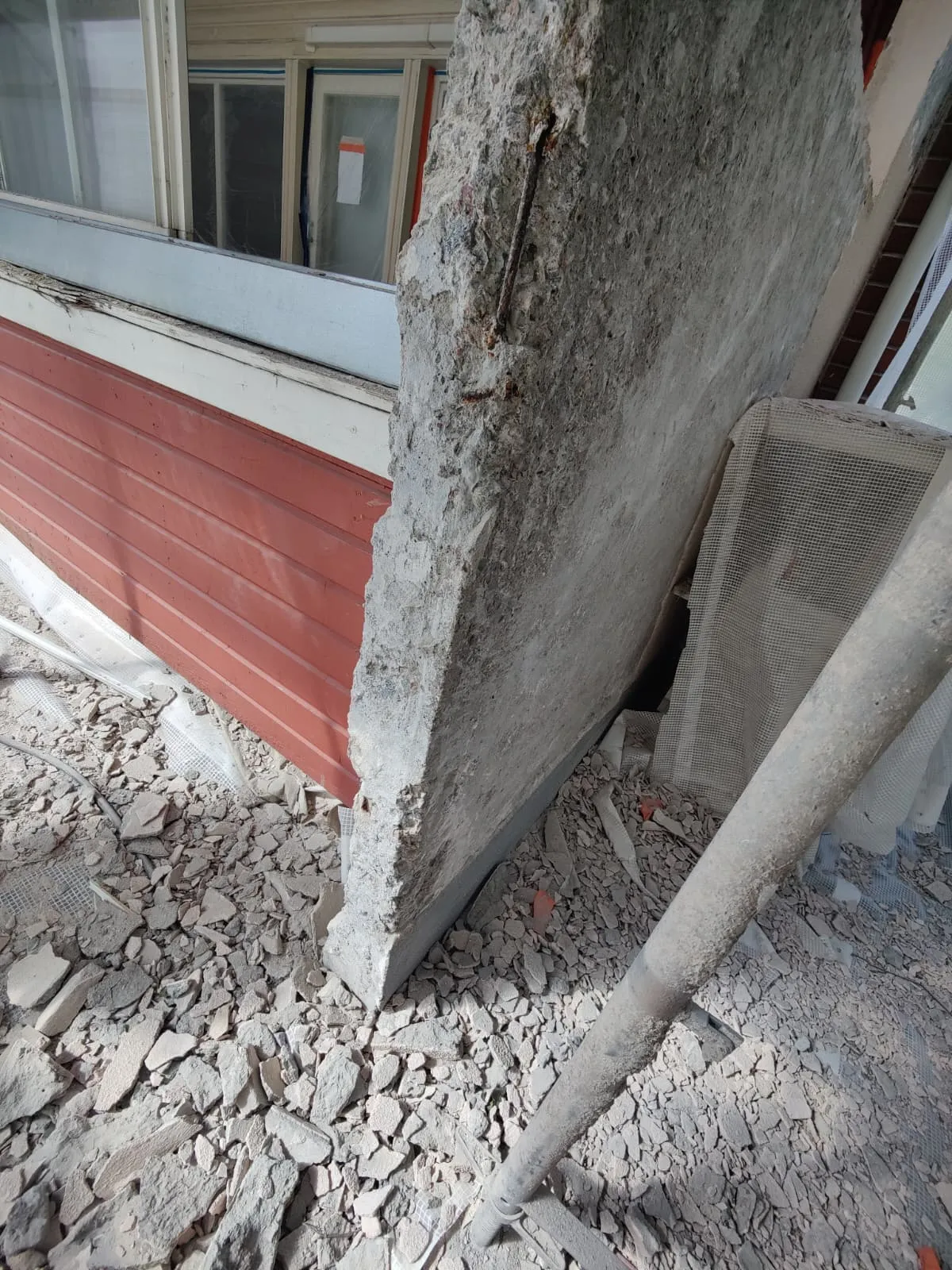 Kuva näyttää betonilaatan, jossa on näkyvissä raudoitusta, sekä punaisen puisen seinän ja ikkunoita. Maassa on betonin sirpaleita ja metalliputki. Valo on kirkas.