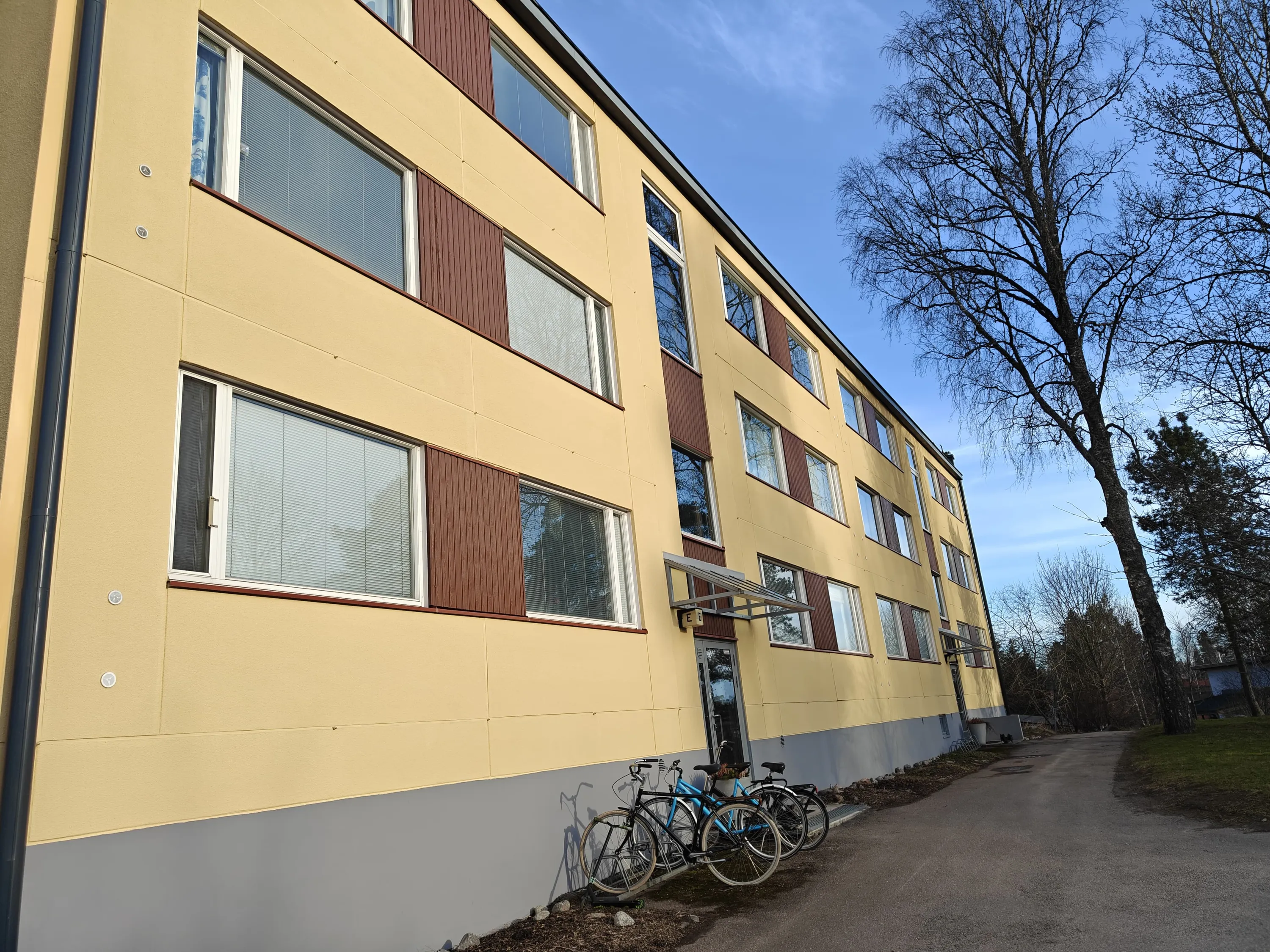 Kolmikerroksinen keltainen rakennus, jossa on ruskeita ikkunaluukkuja, polkupyöriä edessä ja puita taustalla aurinkoisena päivänä.