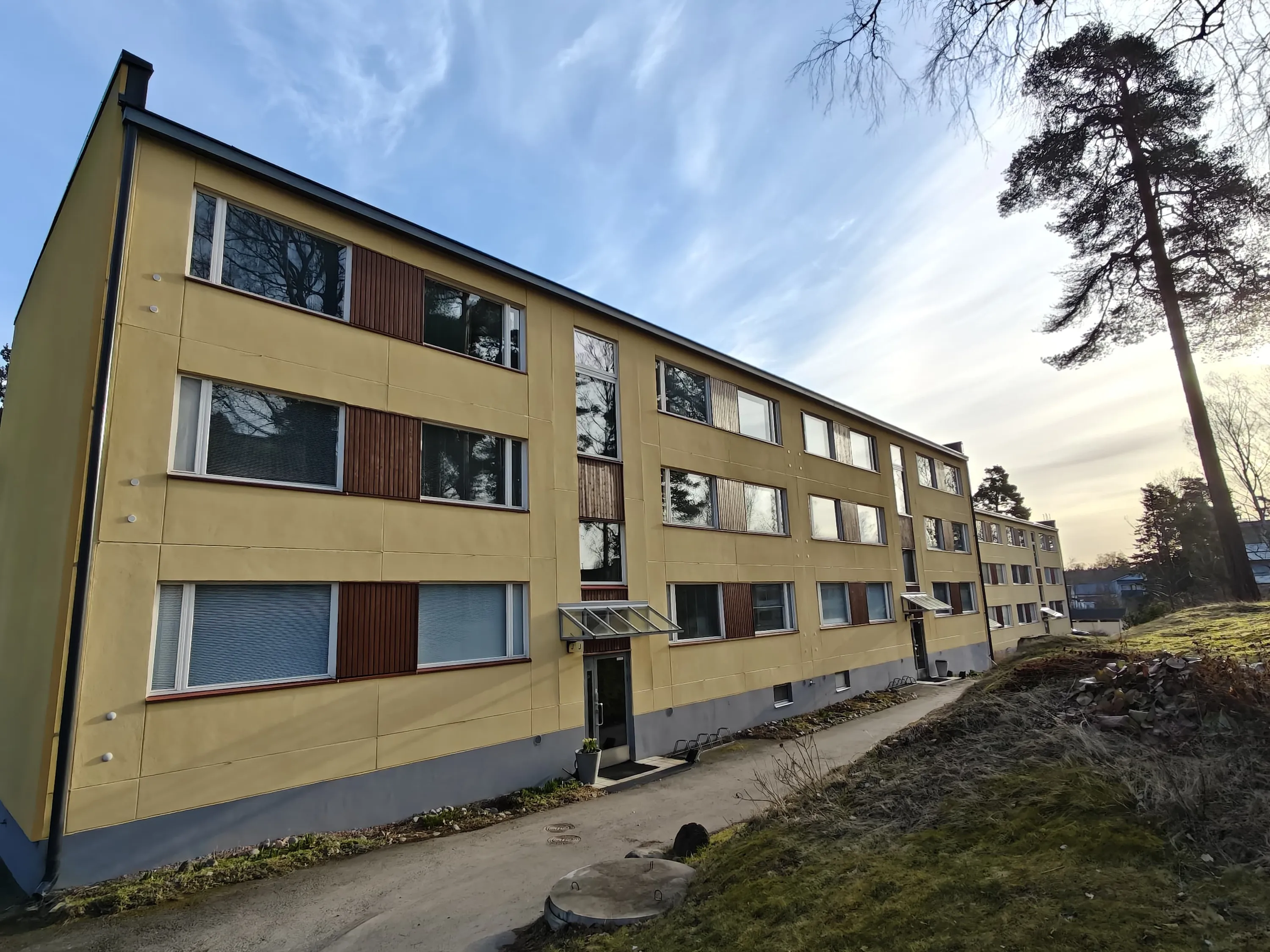 Kolmikerroksinen, keltainen asuintalo, jossa on ruskeat ikkunaluukut ja harmaa pohja, sijaitsee kukkulalla aurinkoisena päivänä.