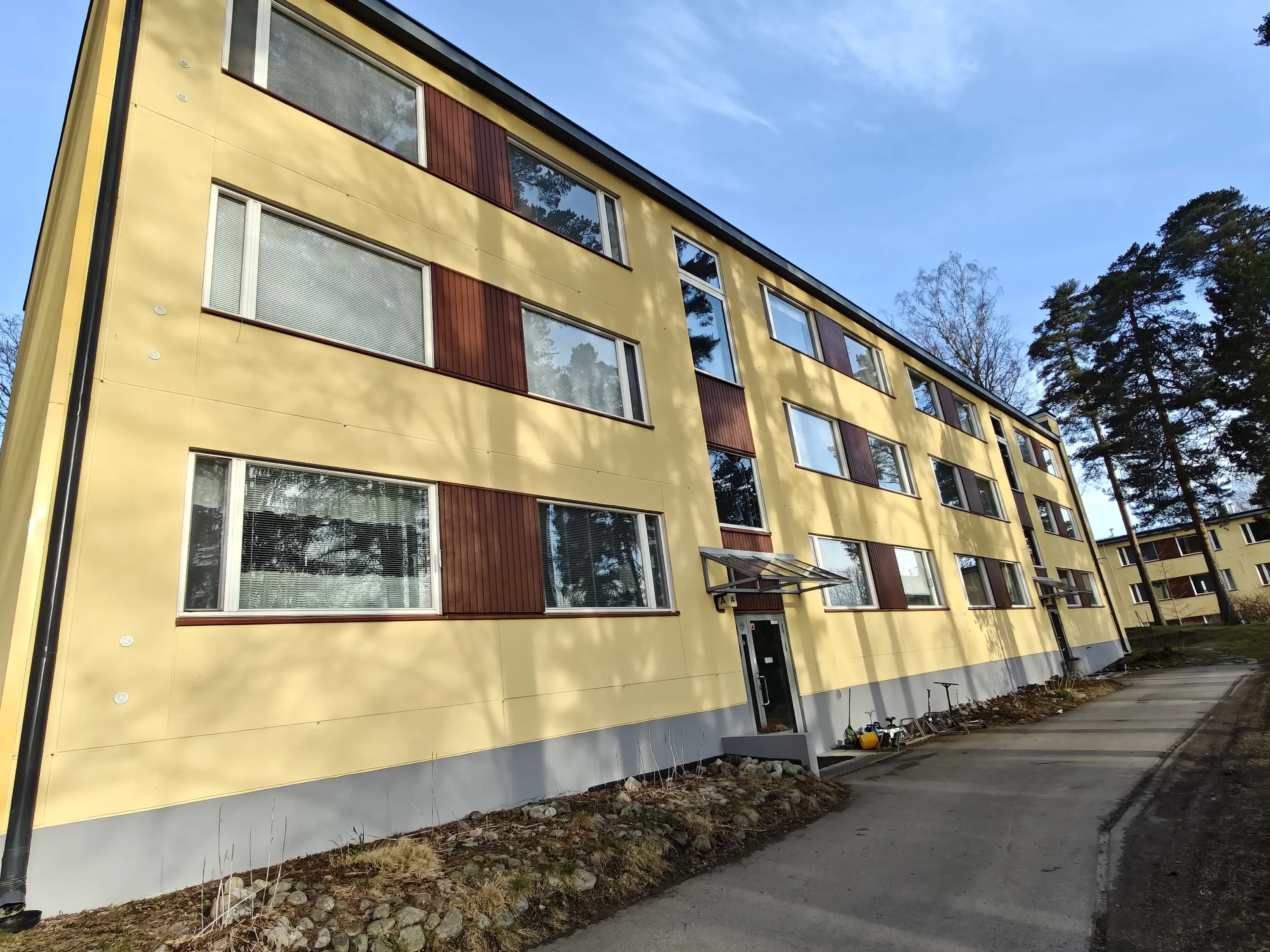 Kaksikerroksinen keltainen asuintalo, jossa on ruskeat ikkunaluukut ja harmaa pohja, valoisassa säässä.