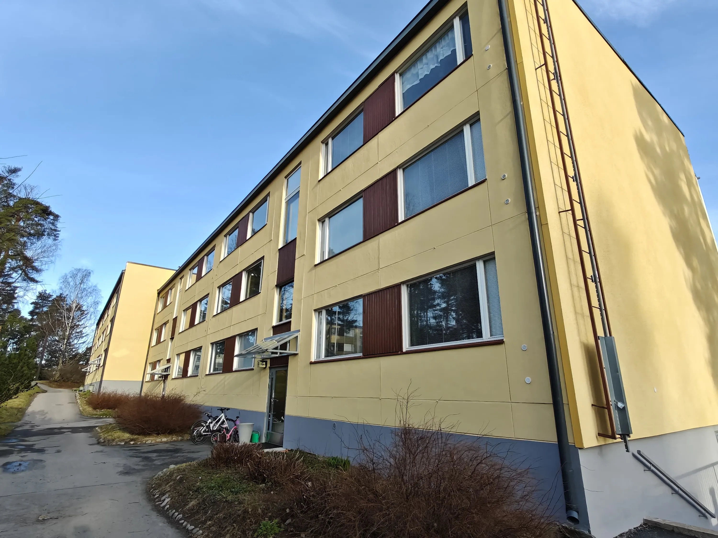 Vaaleankeltainen kerrostalo, jossa on ruskeat ikkunapaneelit ja sininen pohja, aurinkoisena päivänä.