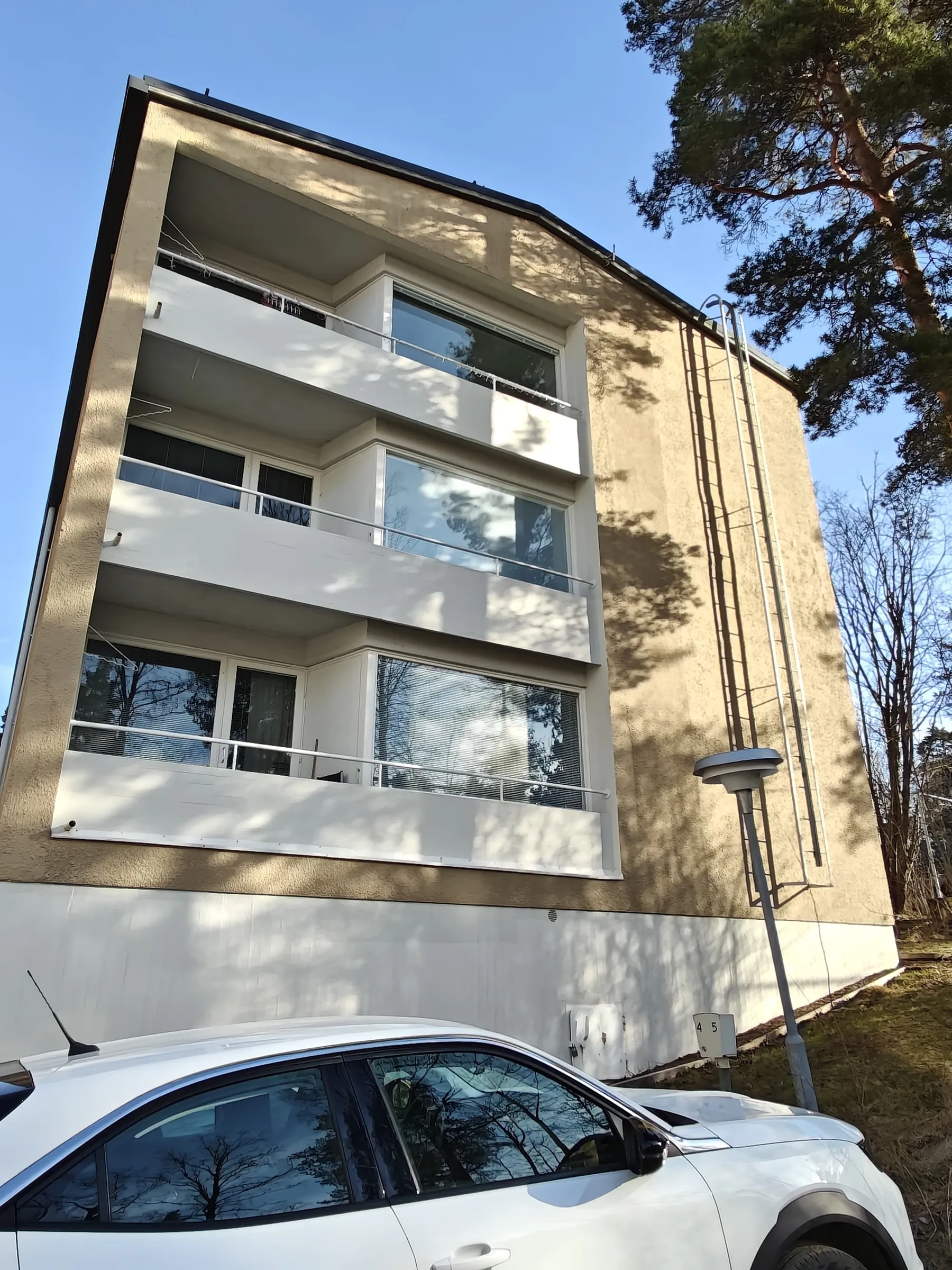 Kolmikerroksinen kerrostalo, jossa on parvekkeet ja valkoinen auto edessä, valaistus on kirkas ja aurinkoinen.