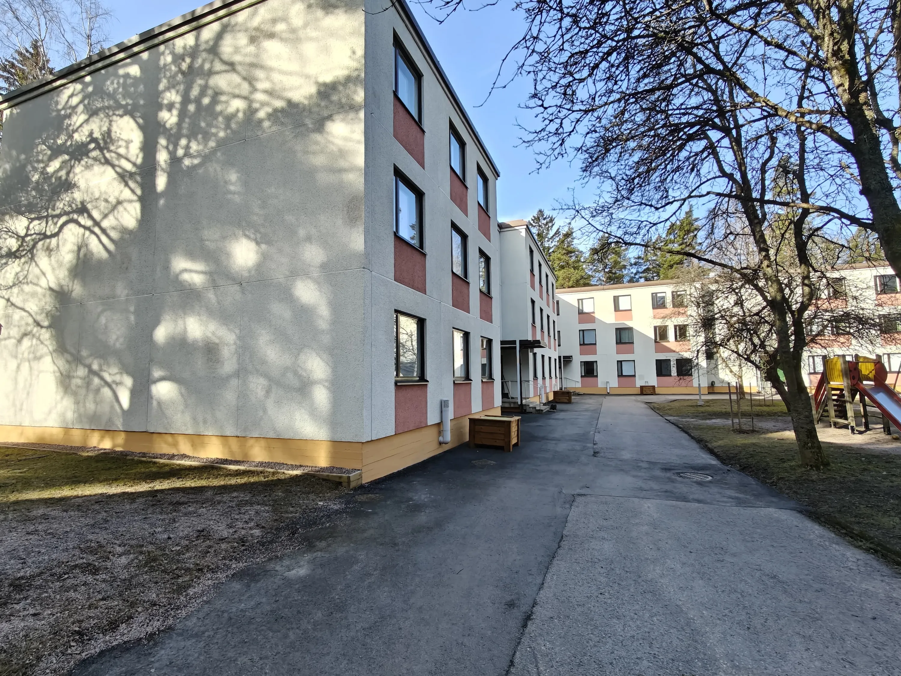 Kolmikerroksinen kerrostalo, jossa on vaaleat seinät, punaiset koristeelliset paneelit ikkunoiden ympärillä ja tummat ikkunat, sekä leikkikenttä ja puita.