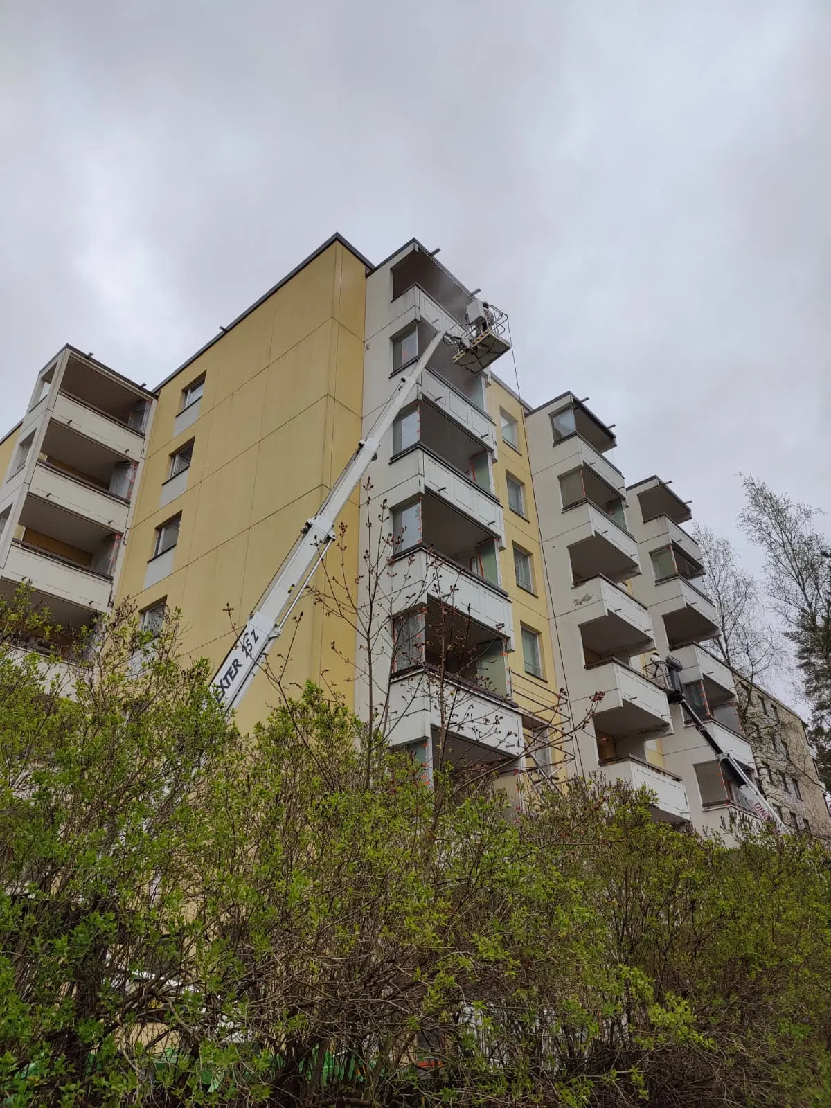 Korkea kerrostalo, jossa on kaksi nosturia, joista toinen on keltaisen seinän edessä ja toinen valkoisen seinän edessä, molemmissa on työntekijöitä korissa.