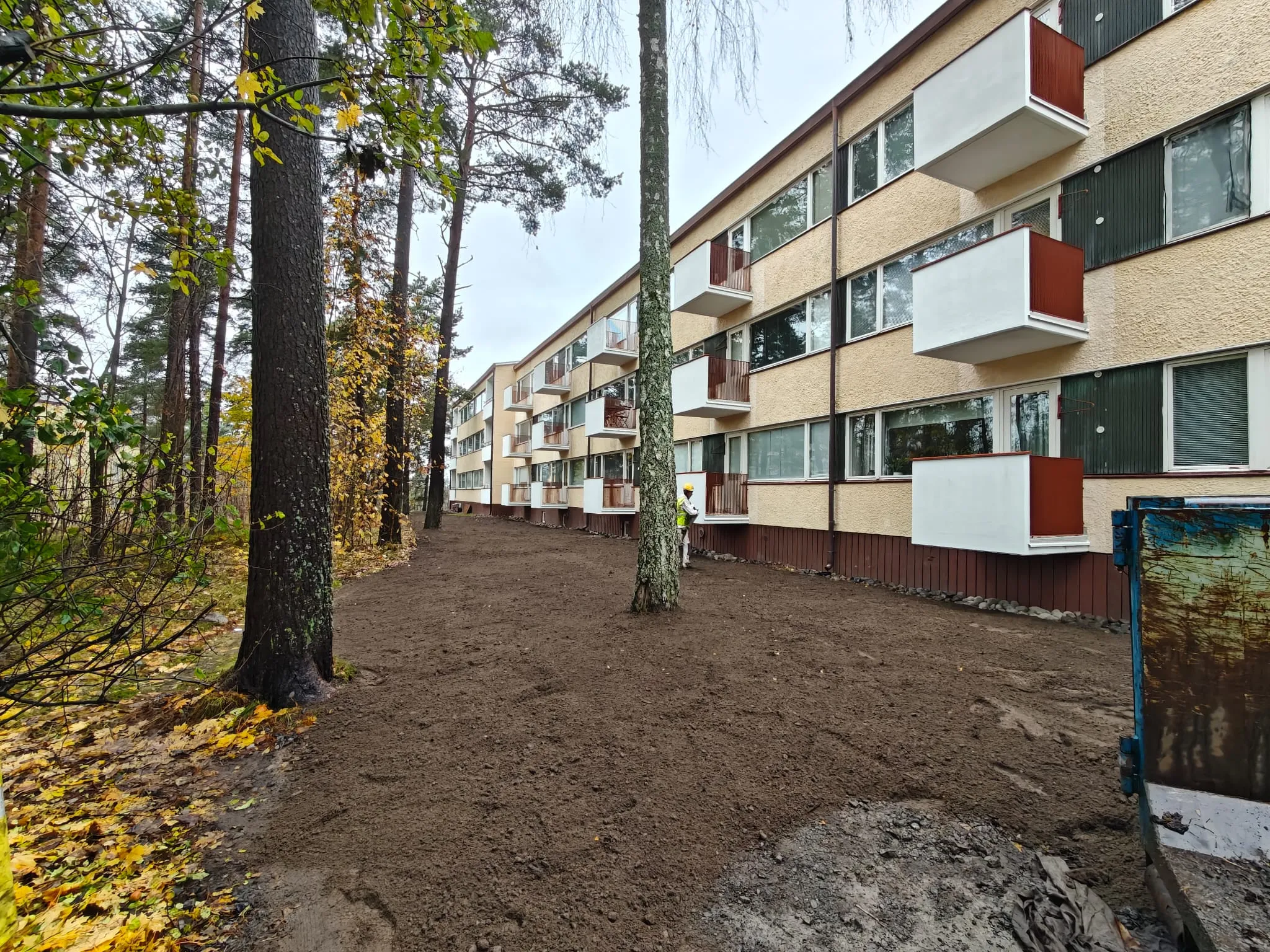 Matala kerrostalo, jossa on parvekkeet ja puita, ruskea maa-alue edessä, harmaa sää.