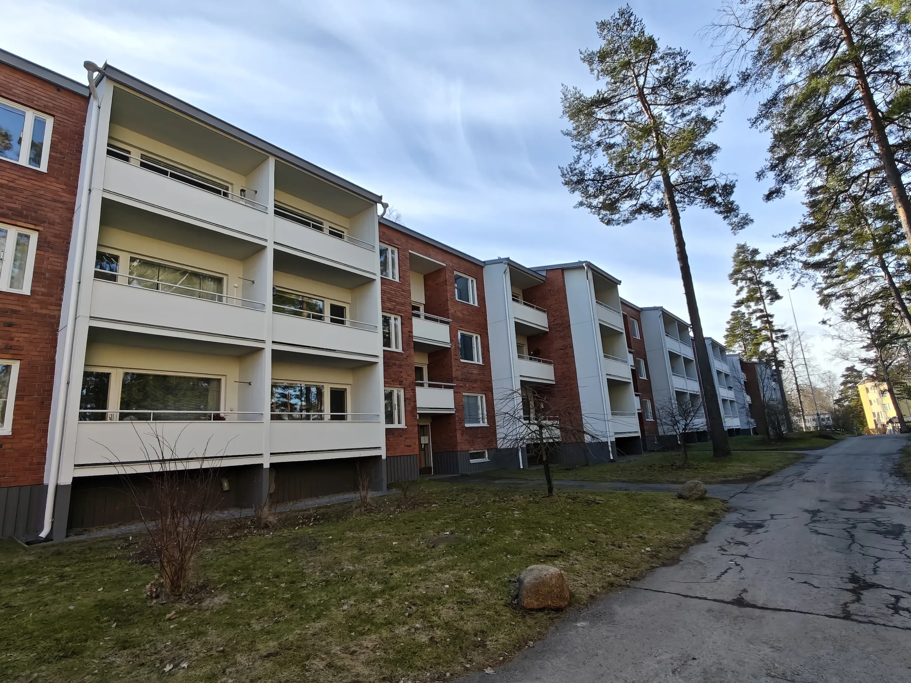 Punatiilinen kerrostalo, jossa on valkoiset parvekkeet ja vihreä nurmikko, seisoo polun vieressä, jota reunustavat korkeat puut sinisen taivaan alla.