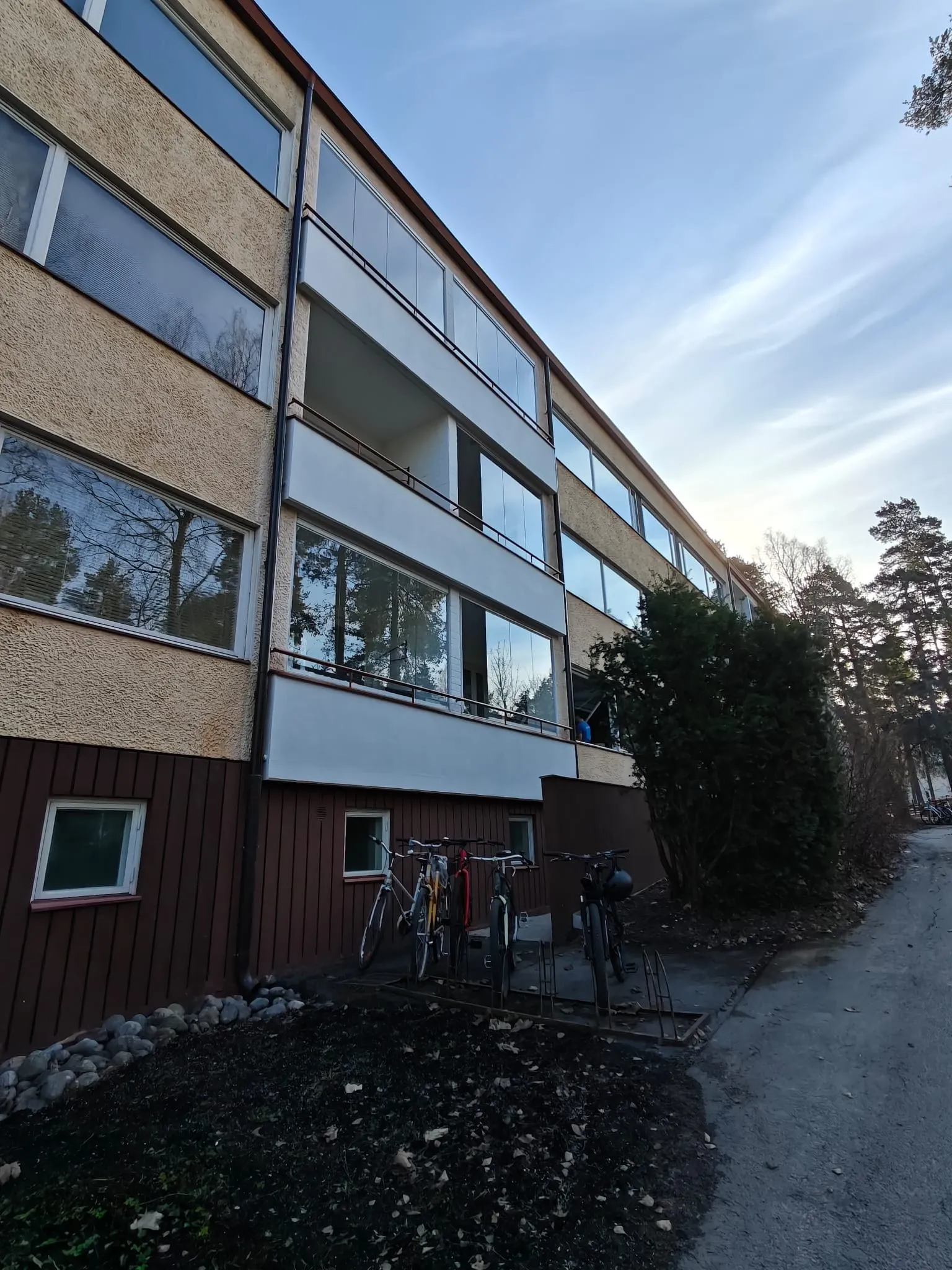 Kolmikerroksinen kerrostalo, jossa on parvekkeet ja pyörätelineitä edessä, valaistus on luonnollinen.