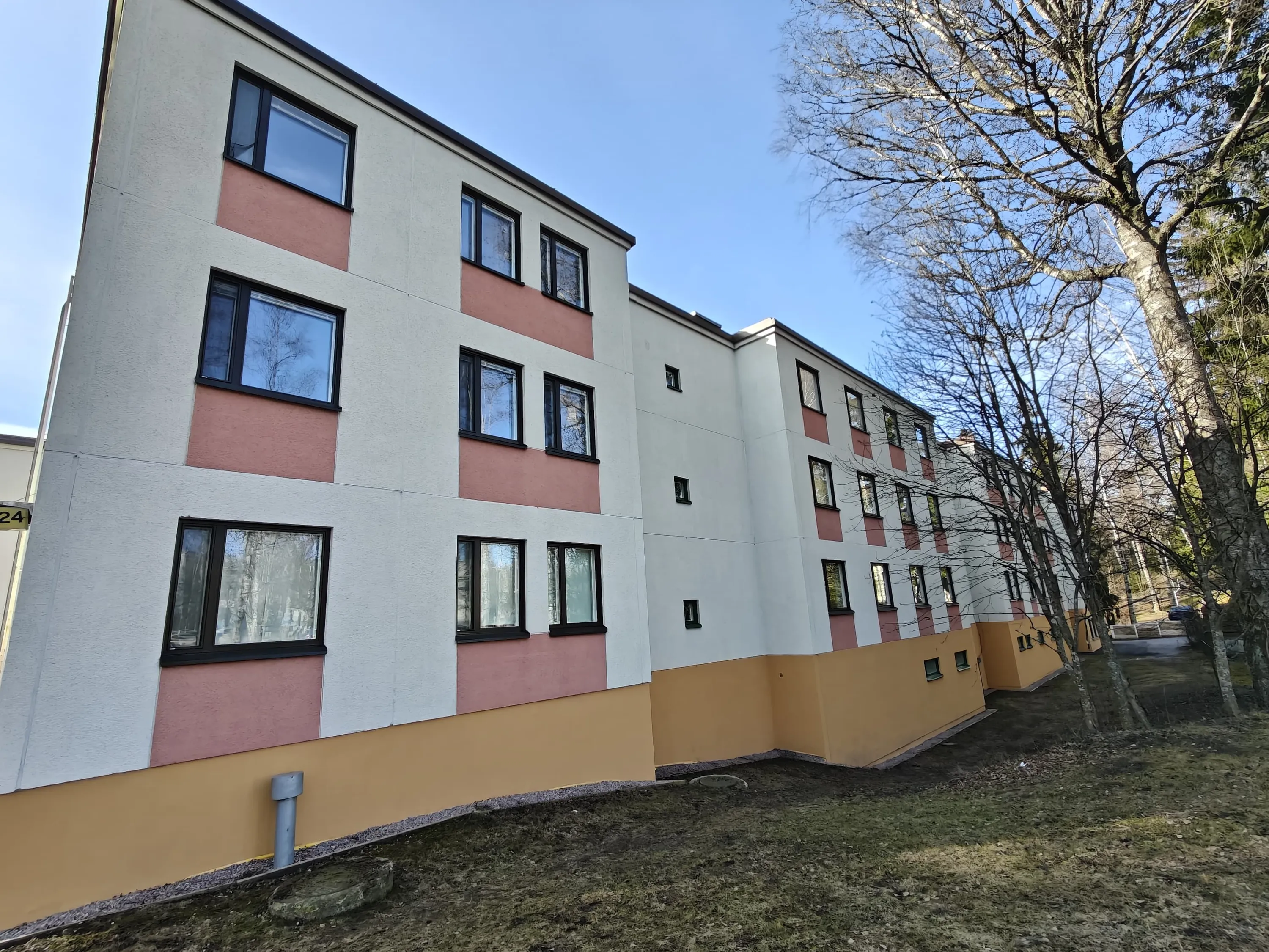 Kolmikerroksinen kerrostalo, jossa on valkoiset seinät, vaaleanpunaiset ja oranssit korostukset, mustat ikkunat ja puita taustalla kirkkaassa säässä.