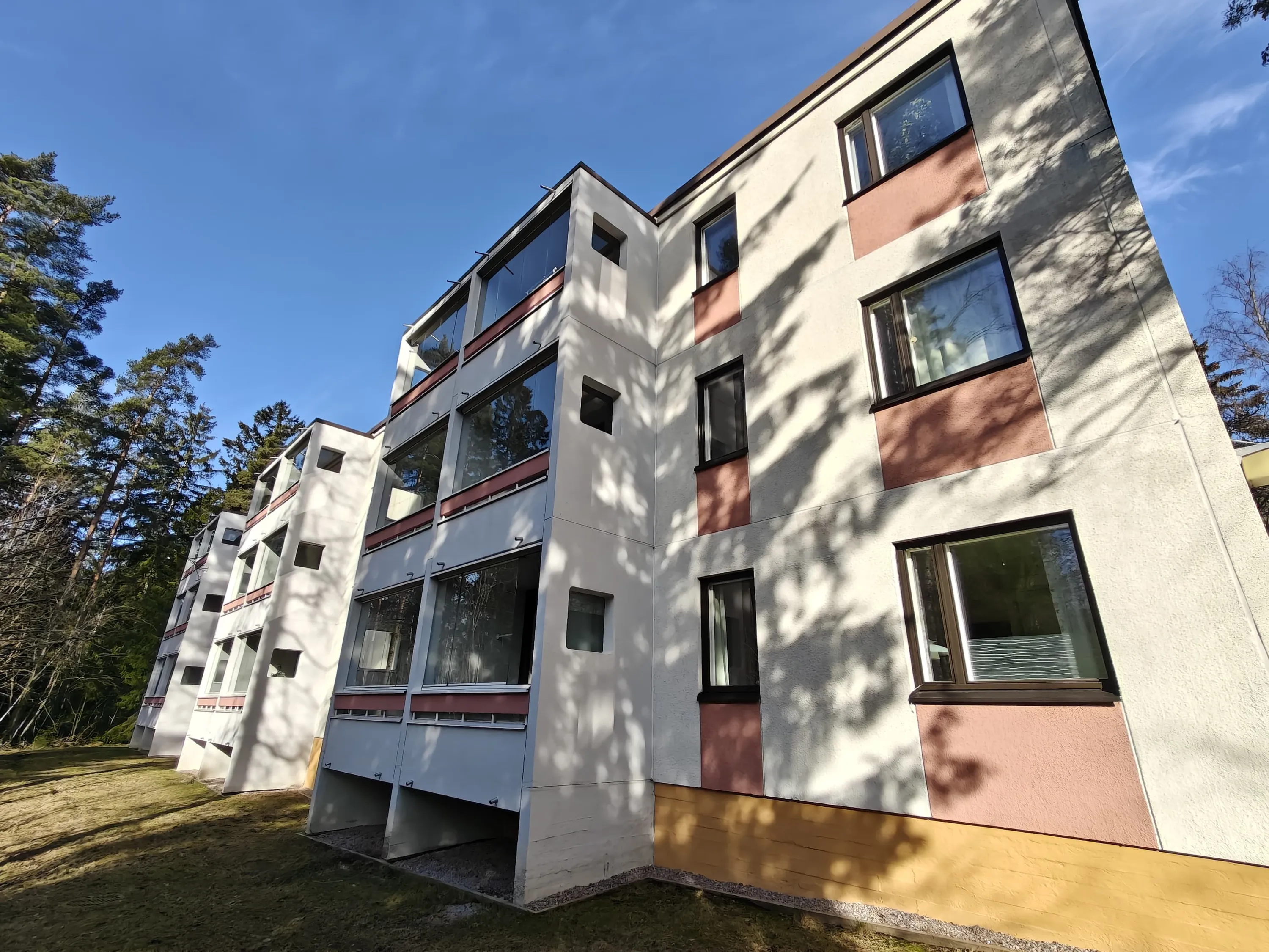 Matala kulma kuvaa kerrostaloa, jossa on valkoiset seinät, ruskeat ikkunat ja parvekkeet, sekä puita ja sininen taivas.