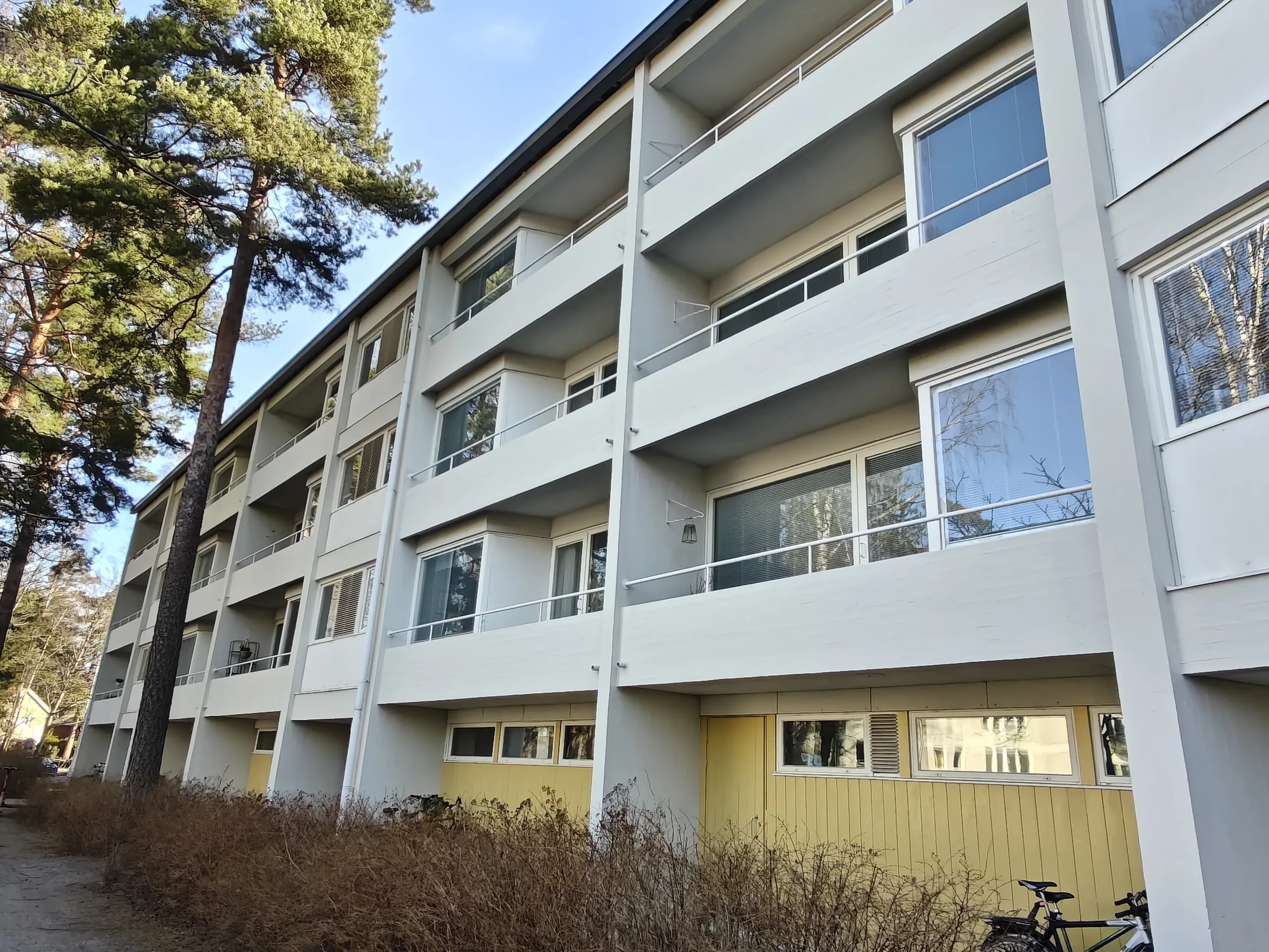 Vaaleanharmaa kerrostalo, jossa on keltaiset pohjakerroksen seinät, parvekkeet ja suuret ikkunat, valoisa sää.