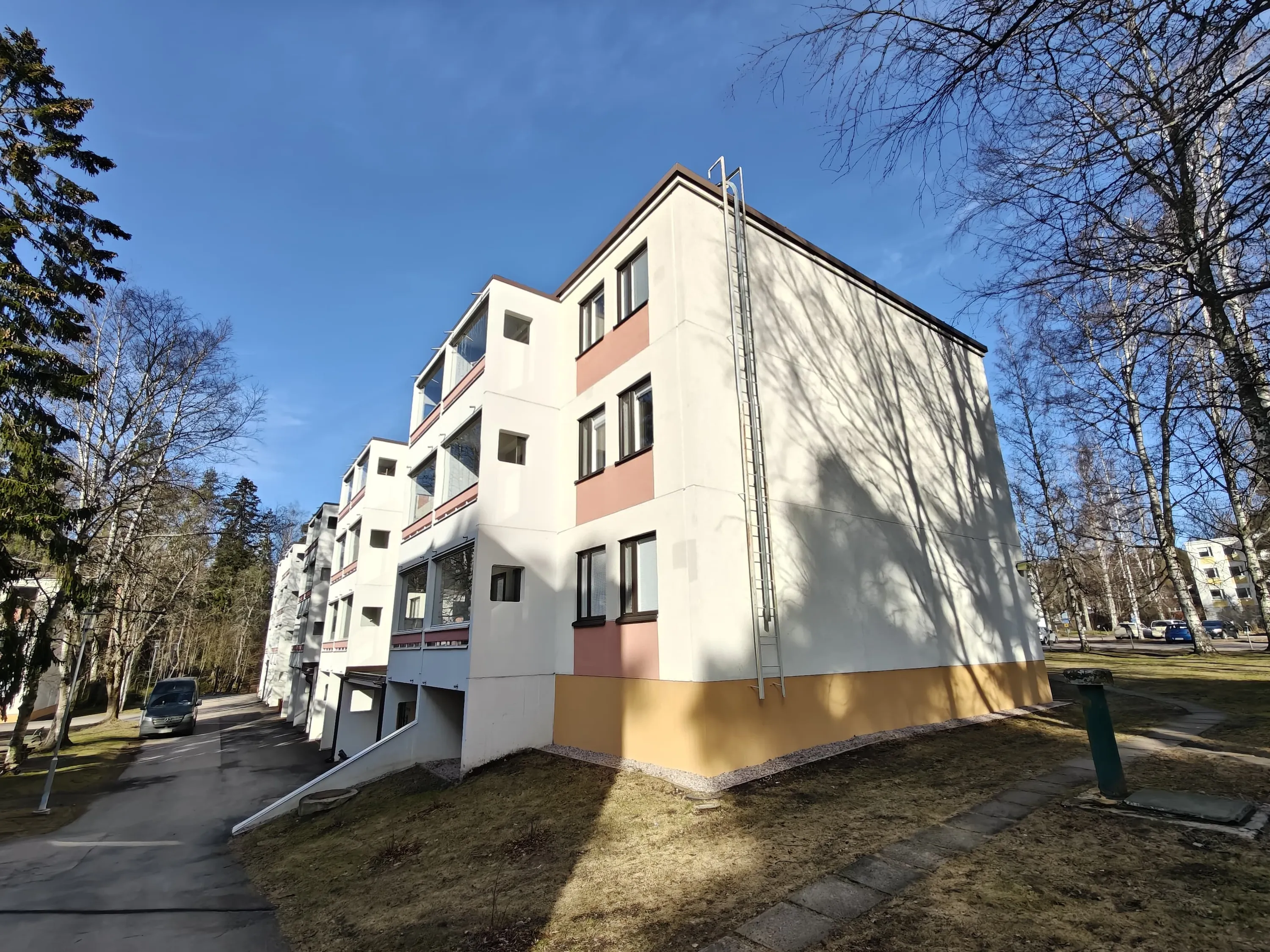 Matala kerrostalo, jossa on valkoiset seinät, ruskeat ikkunakehykset ja keltainen pohja. Rakennuksen seinällä on metallinen palotikas. Taustalla näkyy puita ja sininen taivas.