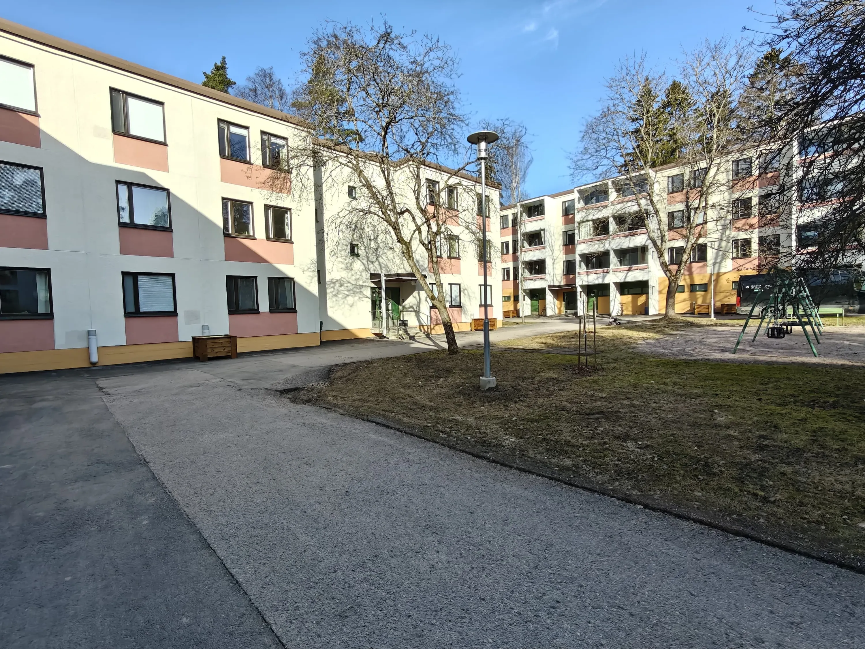 Kolmikerroksisia kerrostaloja pihalla, jossa on puita, nurmikkoa ja leikkivälineitä aurinkoisena päivänä.