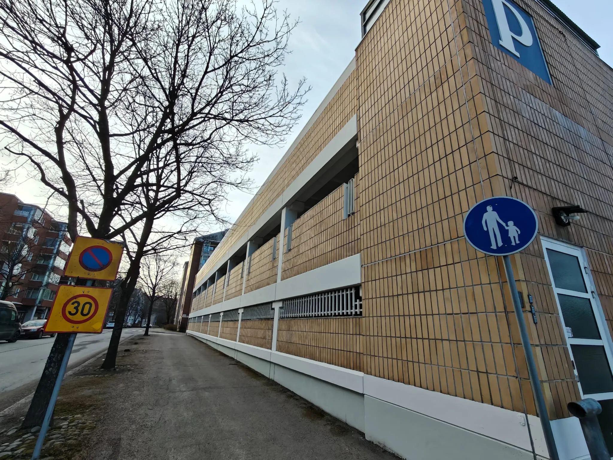 Ulkona on pysäköintitalo, jonka vieressä on jalkakäytävä, jossa on liikennemerkkejä ja puu. Pysäköintitalo on tiilenvärinen ja siinä on sininen P-merkki. Taivas on pilvinen ja valaistus on luonnollinen.