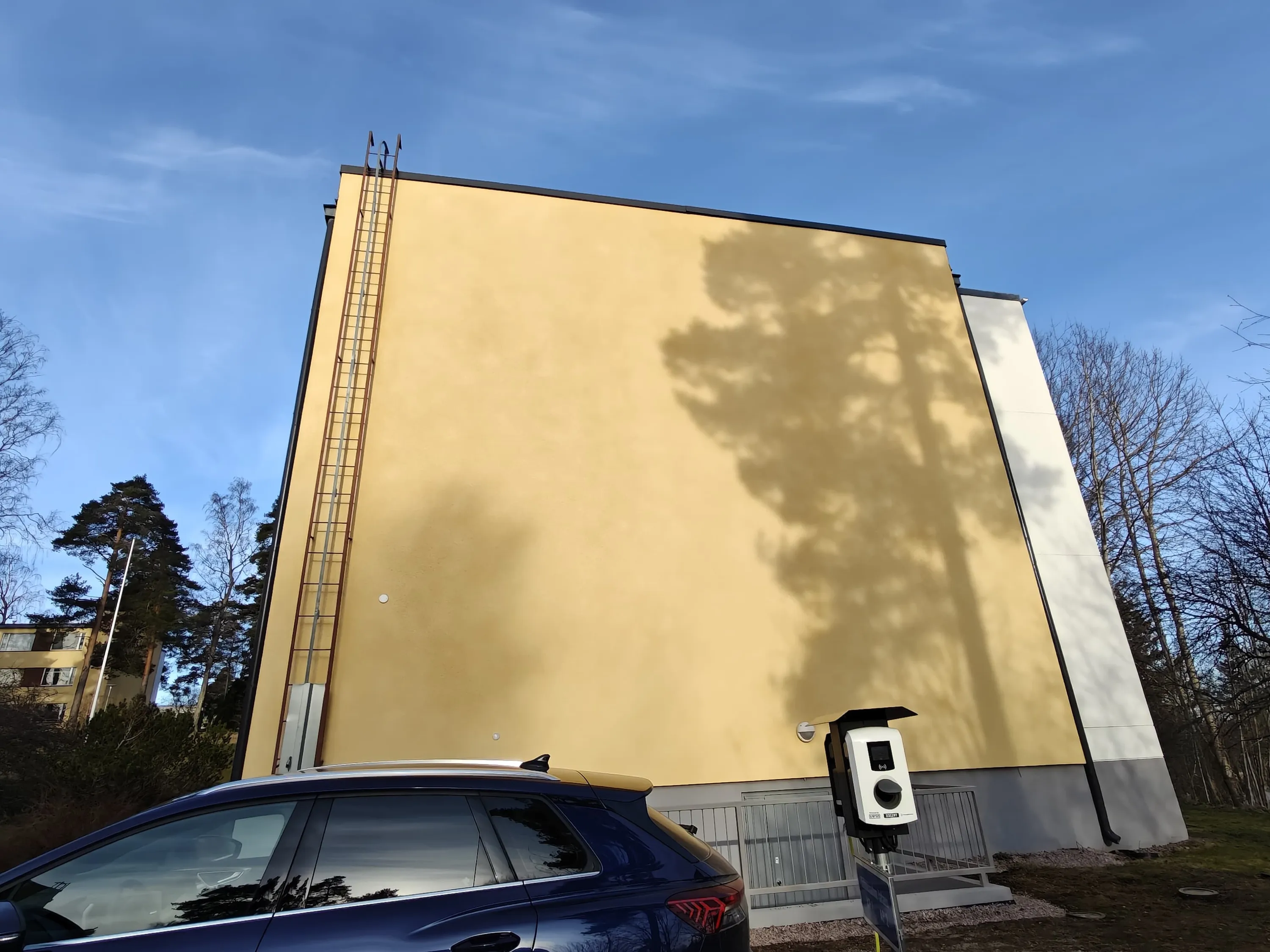 Keltaisen rakennuksen seinällä on pitkät tikkaat ja varjo, vieressä sähköauto ja latausasema aurinkoisena päivänä.