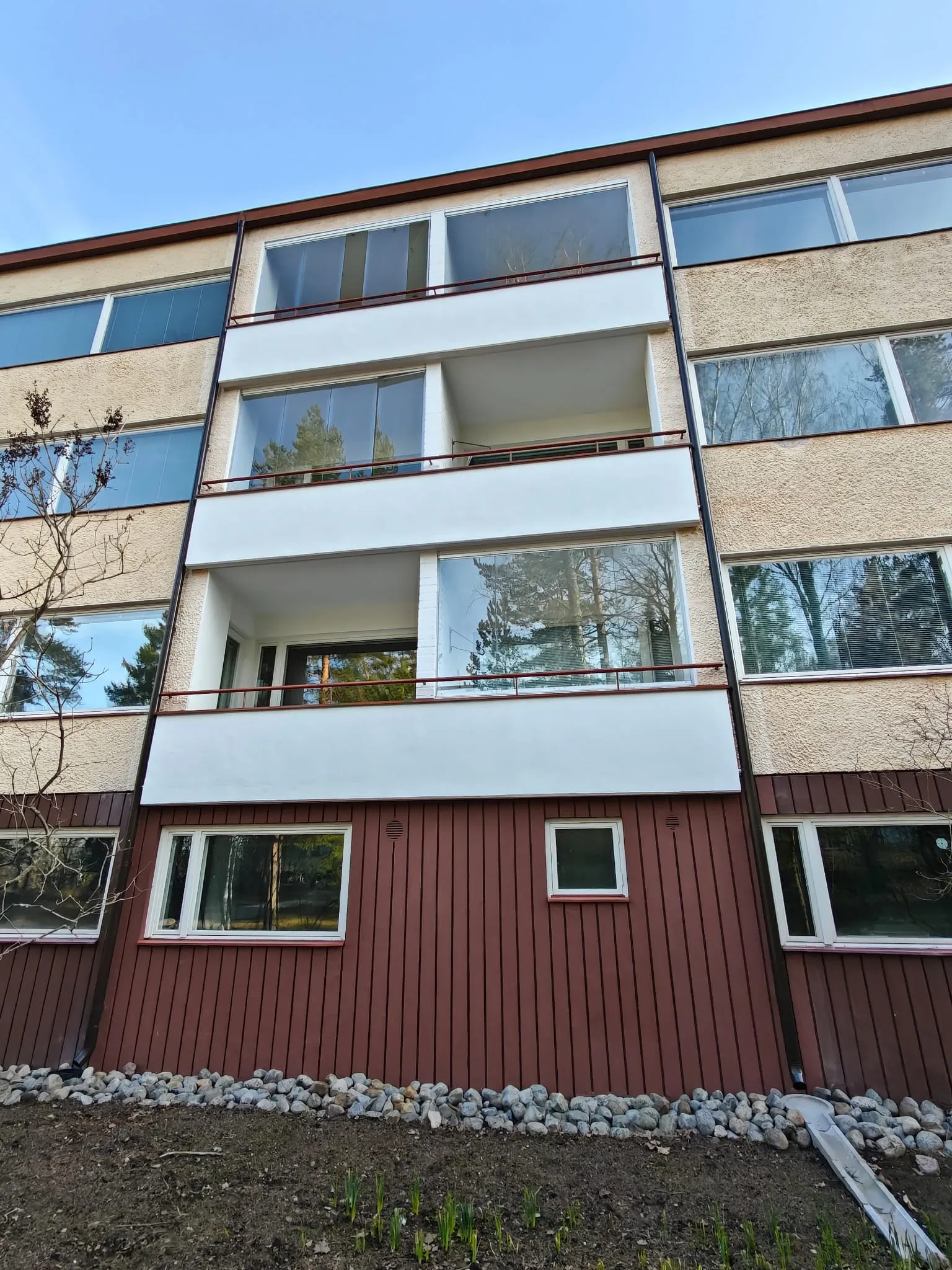 Kerrostalon julkisivu, jossa on ruskea puupanelointi alhaalla, beige rappaus ylhäällä ja lasitetut parvekkeet. Taivas on sininen.