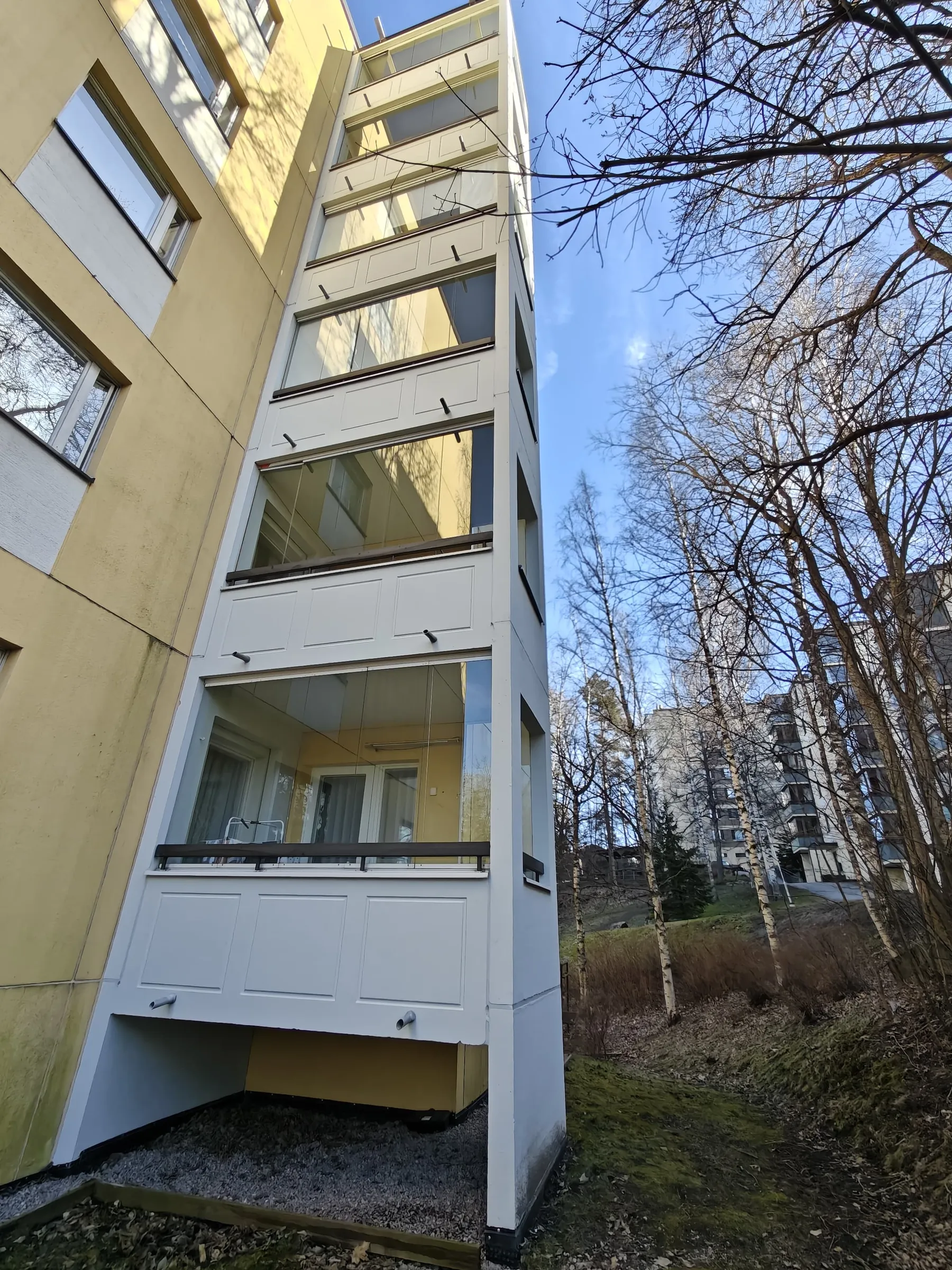 Valokuva rakennuksesta, jossa on lasitetut parvekkeet, otettu alhaalta ylöspäin aurinkoisena päivänä. Rakennus on keltainen ja parvekkeet valkoiset. Taustalla näkyy puita ja sininen taivas.