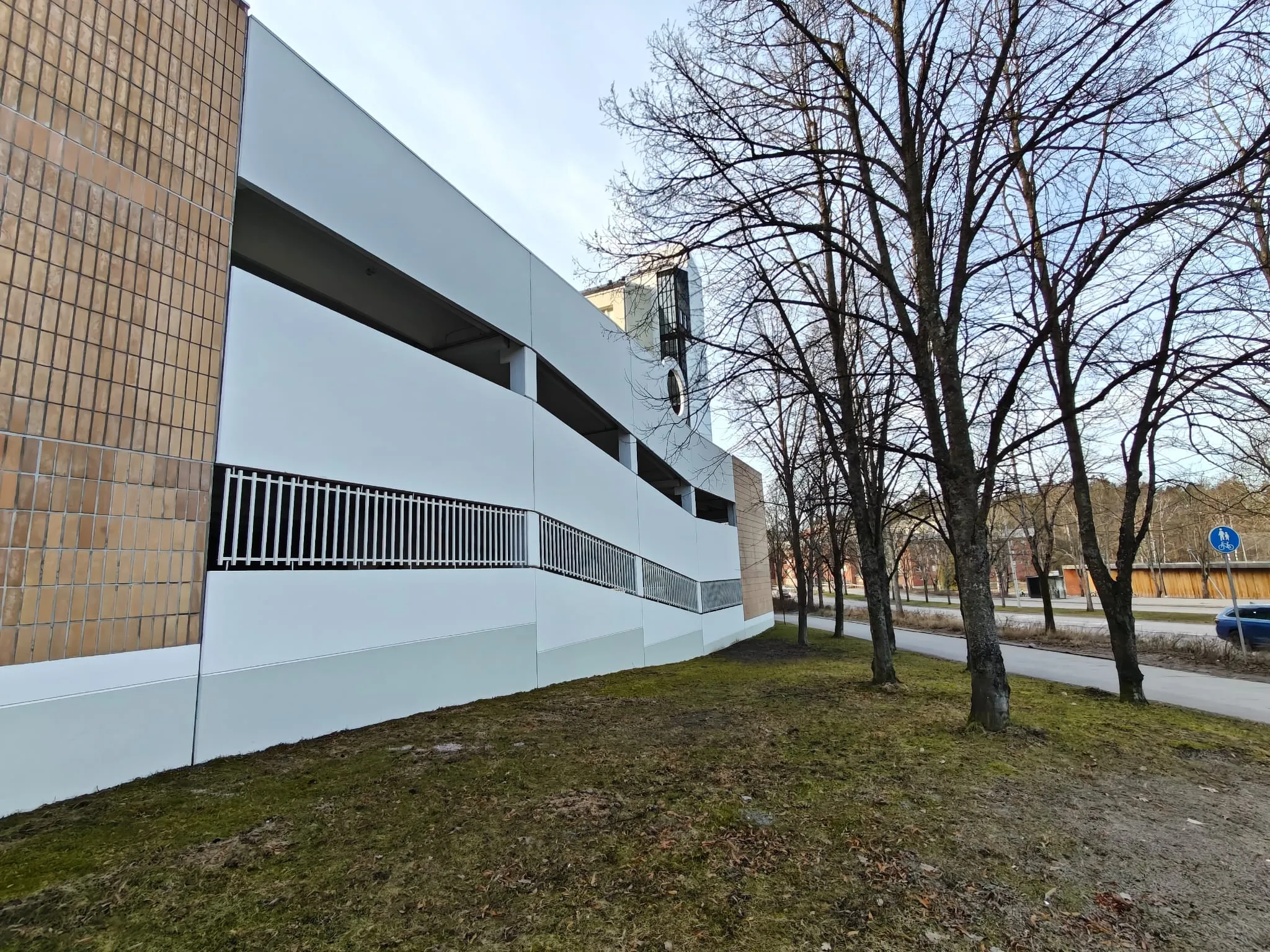 Ulkonäkymä rakennuksesta, jossa on valkoinen julkisivu, ruskea tiiliseinä ja metalliset kaiteet, sekä puita ja nurmikkoa. Valoisa sää.