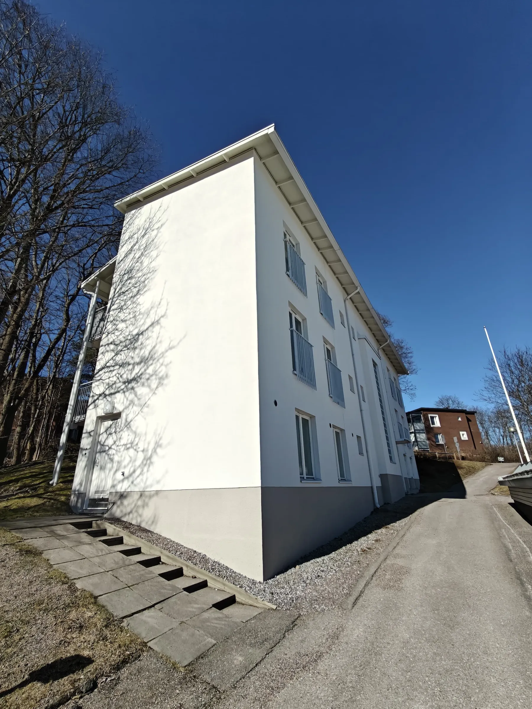 Matala kulma valkoisesta rakennuksesta, jossa on harmaa pohja, sinisen taivaan alla ja puita taustalla.
