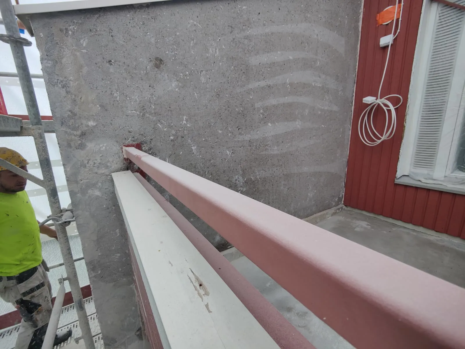 Kuva näyttää rakennustyömaata, jossa on punainen kaide, harmaa seinä ja työmies telineillä. Valo on luonnollinen ja kirkas.