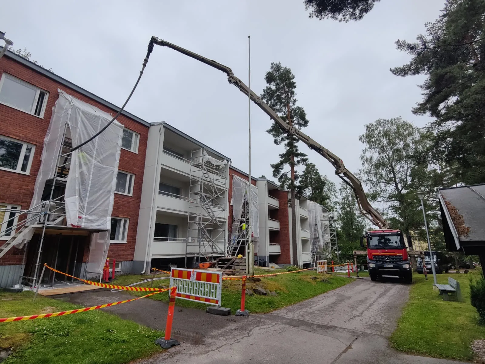 Punatiilinen kerrostalo, jonka julkisivua kunnostetaan, rakennustelineet ja suojamuovit peittävät osan rakennuksesta, betonipumppuauto pumppaa materiaalia ylös, varoitusnauhat ja -merkit ympäröivät työaluetta.