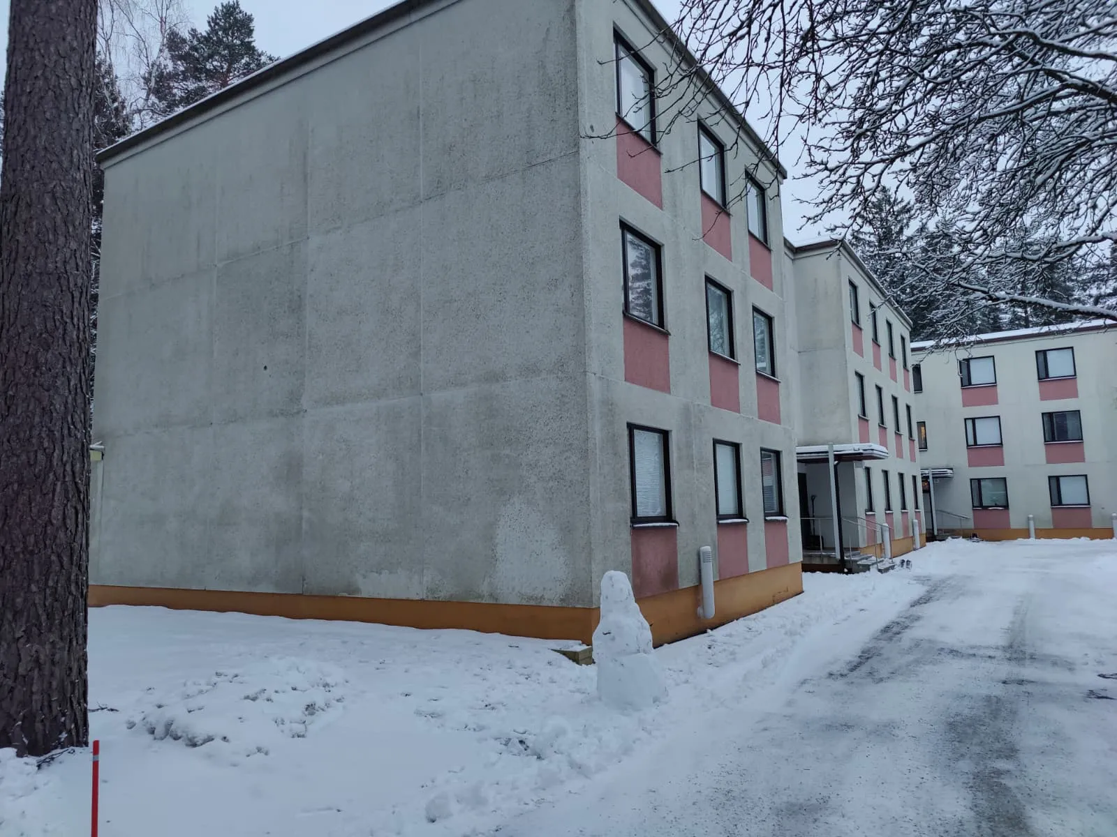 Talvinen maisema, jossa on lumipeitteinen piha ja kolmikerroksisia kerrostaloja, joissa on vaaleanpunaiset koristeet ikkunoiden ympärillä ja pieni lumiukko etualalla.