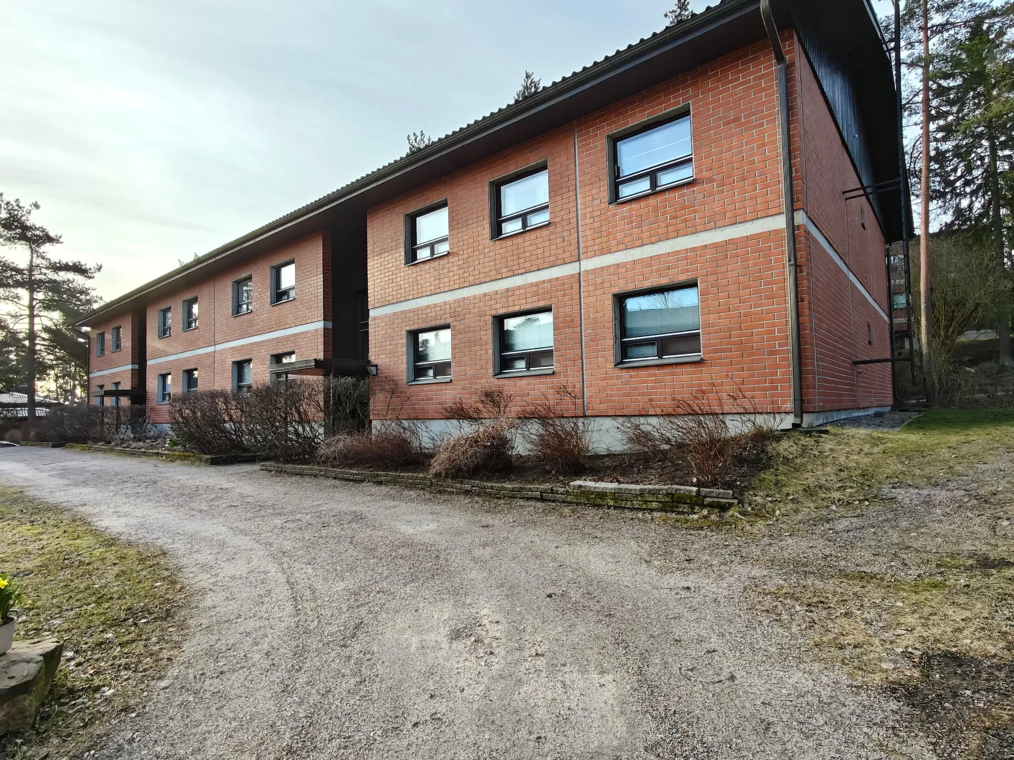 Kaksikerroksinen punatiilitalo, jossa on tummat ikkunat ja harmaa katto, sijaitsee hiekkatien vieressä, jossa on kuivia pensaita ja vihreää ruohoa.