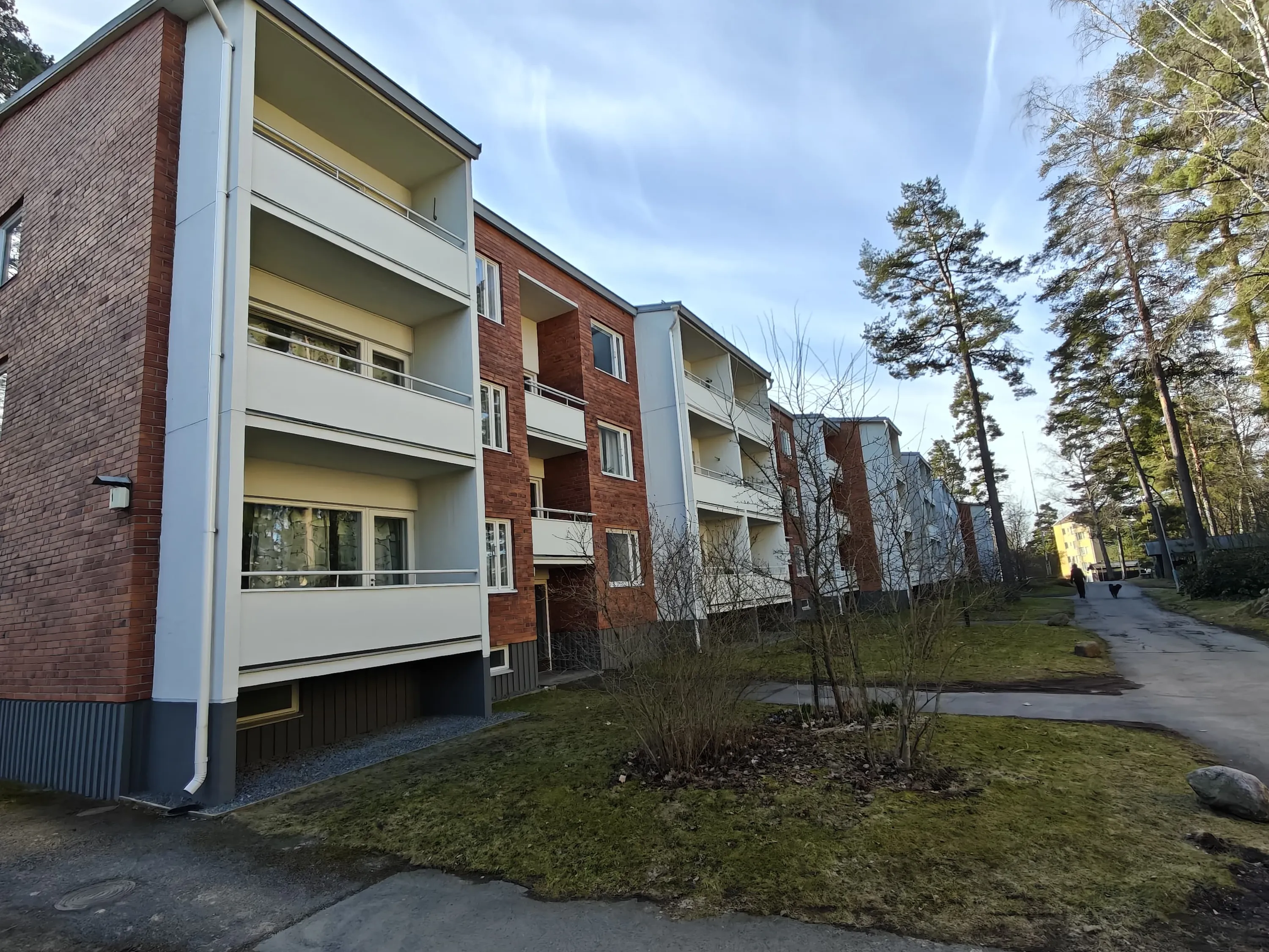 Kolmikerroksinen tiilirakennus, jossa on valkoiset parvekkeet ja vihreä nurmikko, valoisassa säässä.