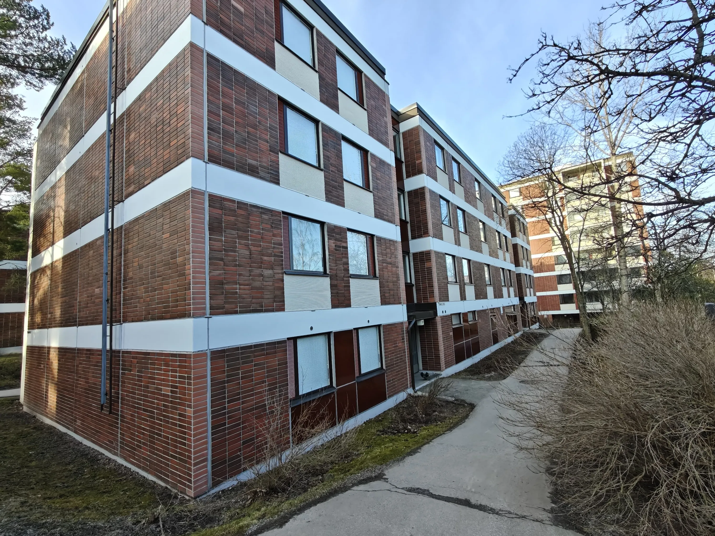 Tiiliverhoiltu kerrostalo, valkoiset vaakasuorat linjat, polku, puita, kirkas valo.