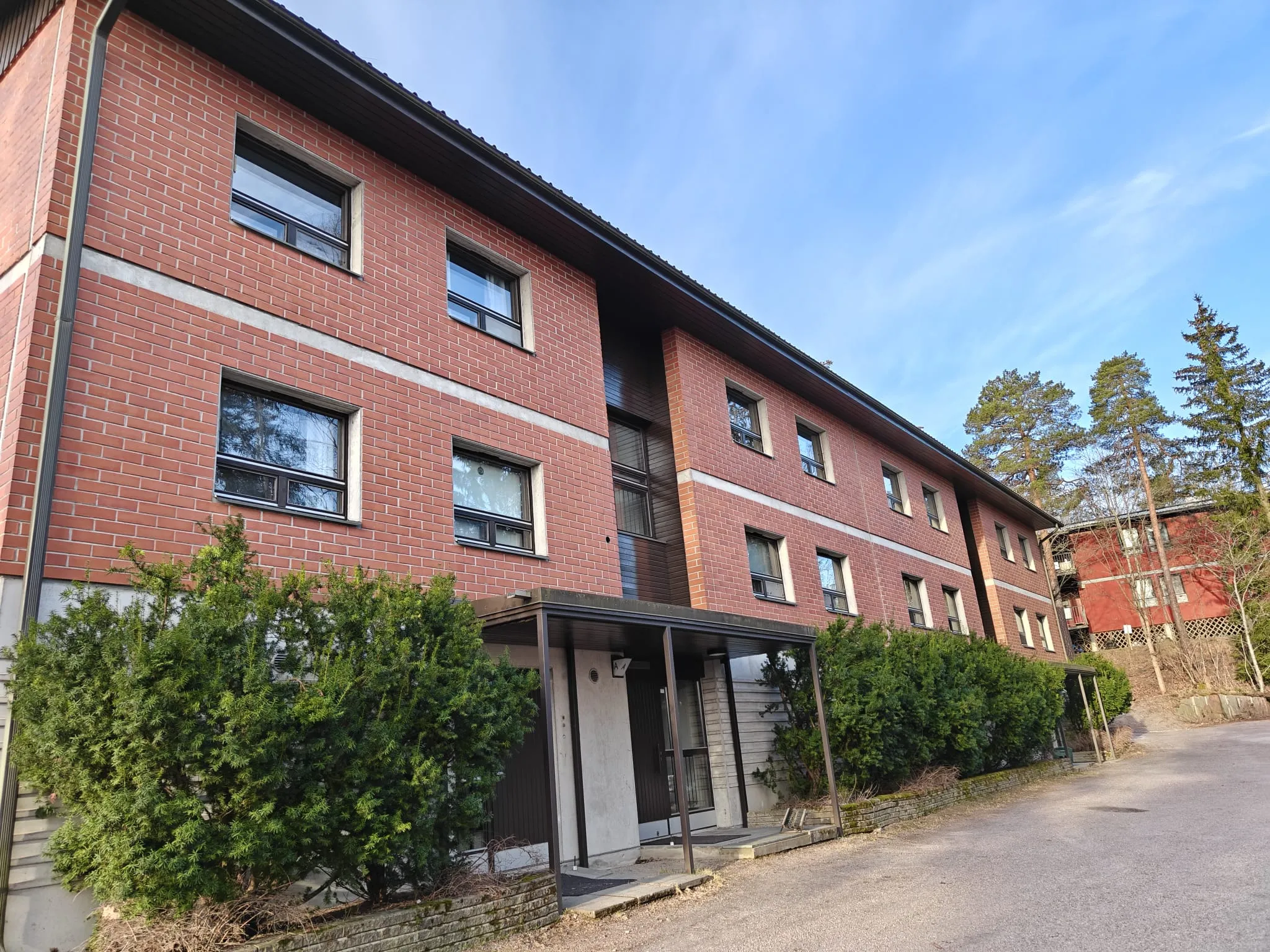 Punatiilinen kerrostalo, jossa on tummat ikkunat ja vihreät pensaat edessä, valoisassa säässä.