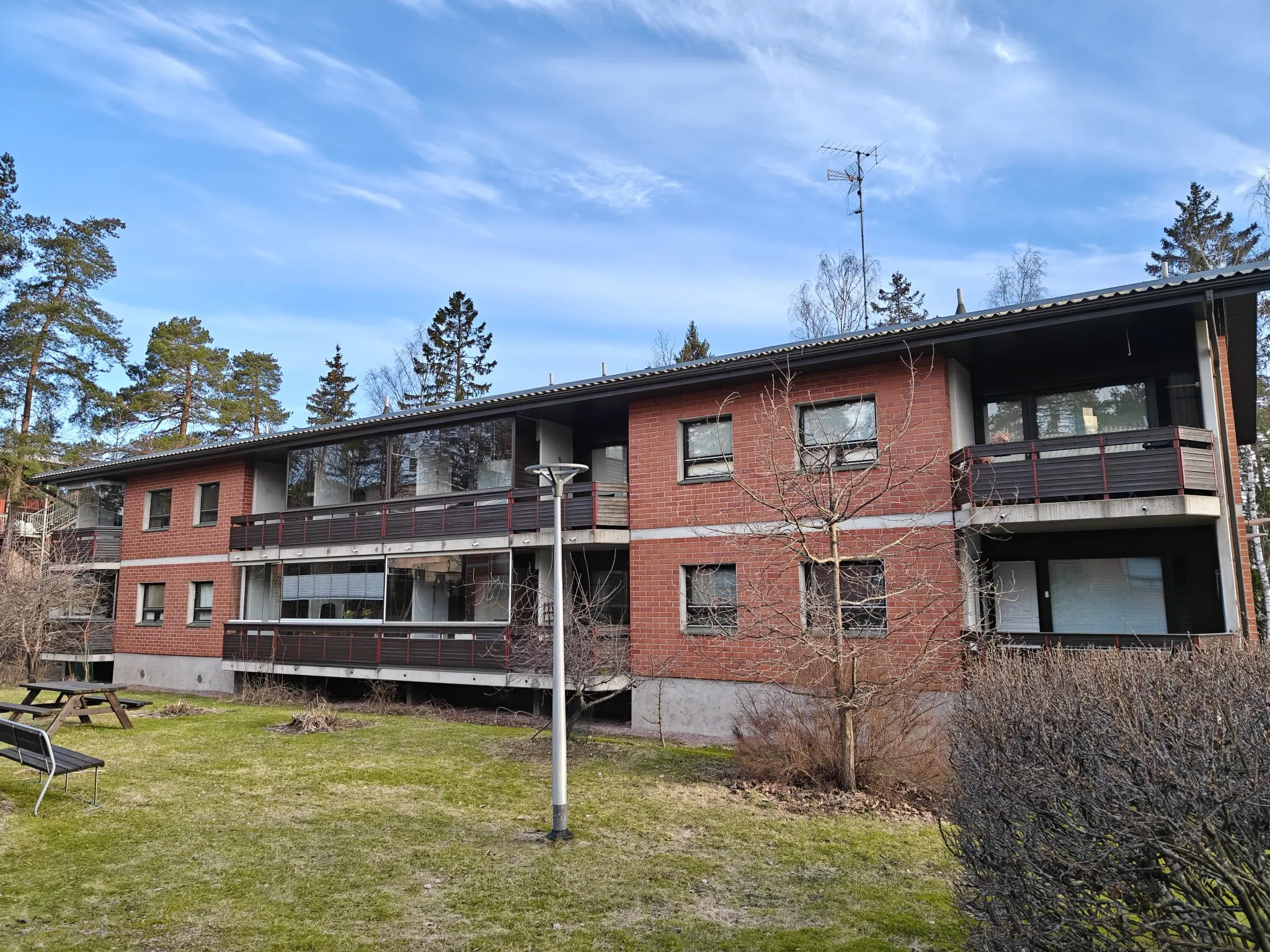 Kolmikerroksinen punatiilitalo, jossa on parvekkeet ja vihreä nurmikko edessä, valaistus on kirkas ja taivas sininen.