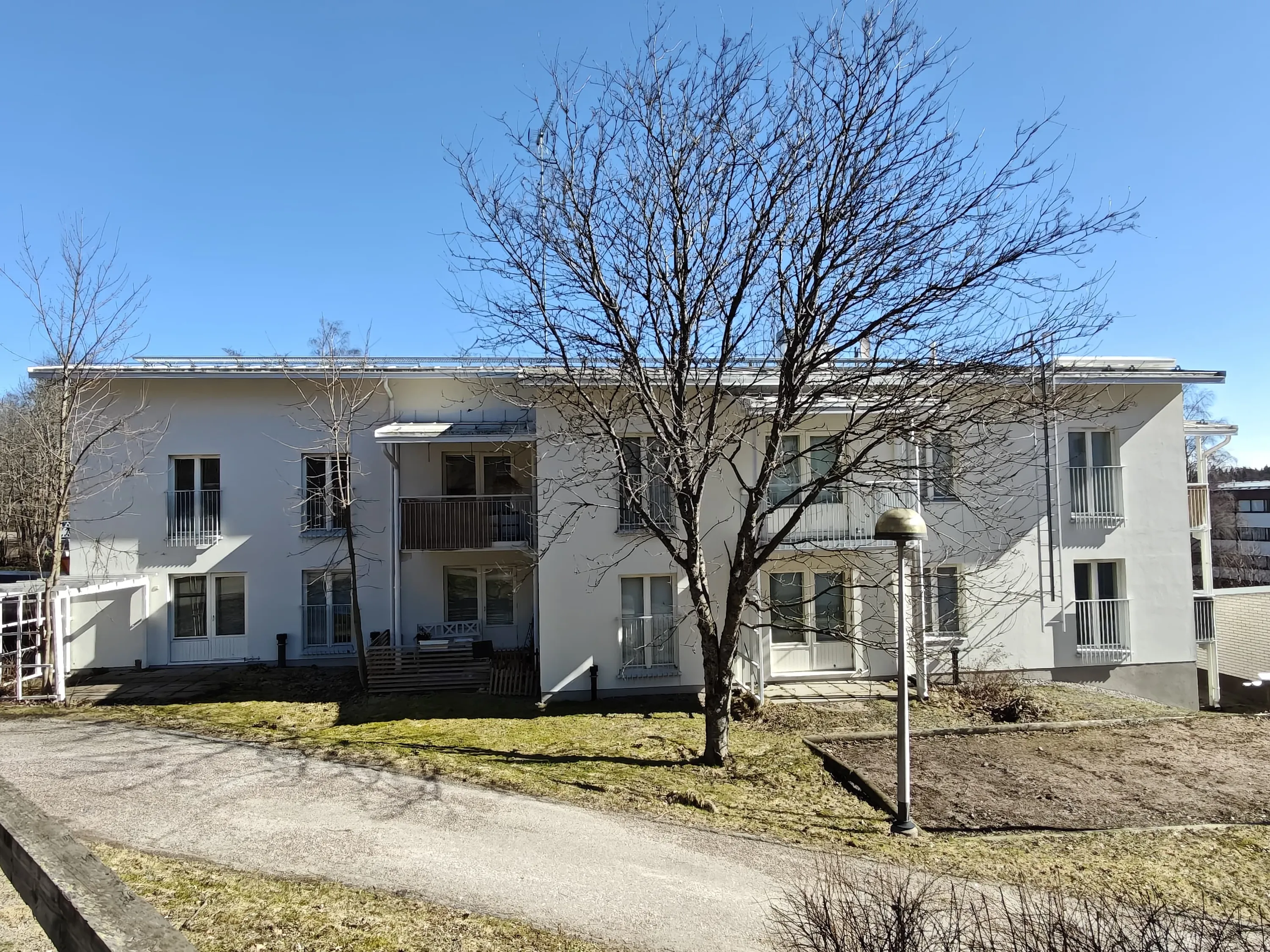 Kaksikerroksinen valkoinen rakennus, jossa on parvekkeita ja suuri puu edessä, aurinkoisena päivänä.